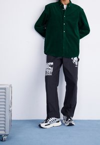 Zielona sztruksowa koszula na guziki, czarne spodnie z białym nadrukiem graficznym oraz czarno-białe buty sportowe. Stoi na jednolitym tle.