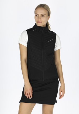 Zwarte mouwloze vest met een hoge kraag, gewatteerde textuur, twee zijzakken en reflecterende accenten. Draagt over een wit T-shirt met korte mouwen.