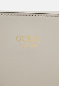 Beige fusk läder plånbok med präglad guld "GUESS" logotyp och "EST 1981." Slät yta med fin sömnad längs kanterna.