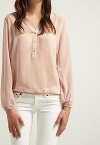 Blusa rosa chiaro con maniche lunghe, scollo a V e dettagli con bottoni. Presenta un sottile motivo a pois e un orlo elastico. Abbinata a pantaloni bianchi.