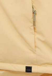 Gros plan d’un tissu jaune pâle avec un nœud de cordon noir et blanc et une petite étiquette noire de la marque Pull&Bear en bas.
