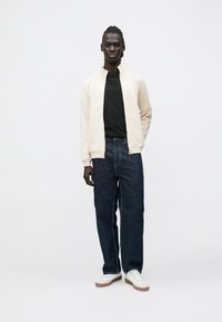 Lys beige bomberjakke over en svart t-skjorte, kombinert med løse mørke denimjeans og hvite joggesko med brune såler. Enkel design, casual passform.