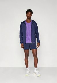 Marineblauw lichtgewicht jack, paarse sportshirt en marineblauwe korte broek. Model draagt witte sneakers; kenmerken zijn onder meer ritsen en logo-accenten.