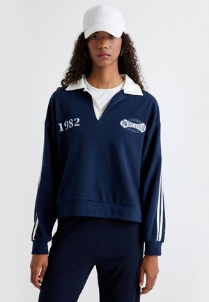 Sweat-shirt bleu marine avec col et rayures blanches, arborant le texte "1982" et le logo "PACIFIC SWEATER" dans un design sportif classique.