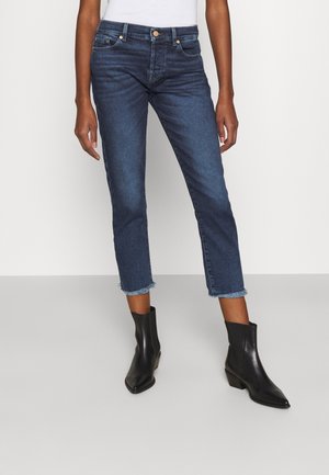 Vaqueros cortos de color azul oscuro con bajos deshilachados, combinados con botines negros y una blusa blanca, mostrados de la cintura a los pies.