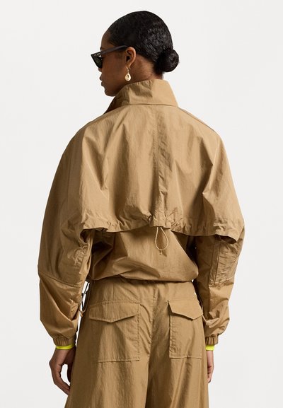 Brūns viegls mētelis ar pārmērīgu piegriezumu, savilktu jostasvietu, elastīgām piedurknēm un augstu apkakli. Saskaņots ar atbilstošām biksēm un stilīgām saules brillēm.