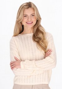 Licht crèmekleurige trui met een gestructureerd opengebreid patroon, voorzien van een ronde hals en geribbelde manchetten. Model poseert met gekruiste armen.