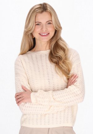 Licht crèmekleurige trui met een gestructureerd opengebreid patroon, voorzien van een ronde hals en geribbelde manchetten. Model poseert met gekruiste armen.