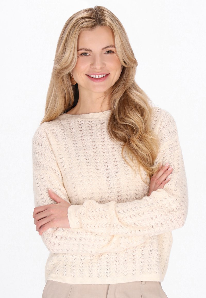 Licht crèmekleurige trui met een gestructureerd opengebreid patroon, voorzien van een ronde hals en geribbelde manchetten. Model poseert met gekruiste armen.