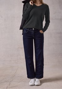 Femme portant un haut à manches longues gris foncé, un jean large bleu foncé, des baskets blanches, tenant une veste noire sur l'épaule, debout sur un sol texturé.