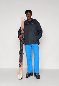 Uomo con giacca nera, pantaloni da sci blu e scarponi da sci, che tiene un paio di sci, con occhiali da sci sulla fronte, in piedi su uno sfondo neutro.