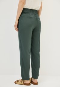 Dark green, lightweight pants with a smocked waistband, tapered leg design, and back pocket detail. Worn with brown woven sandals.