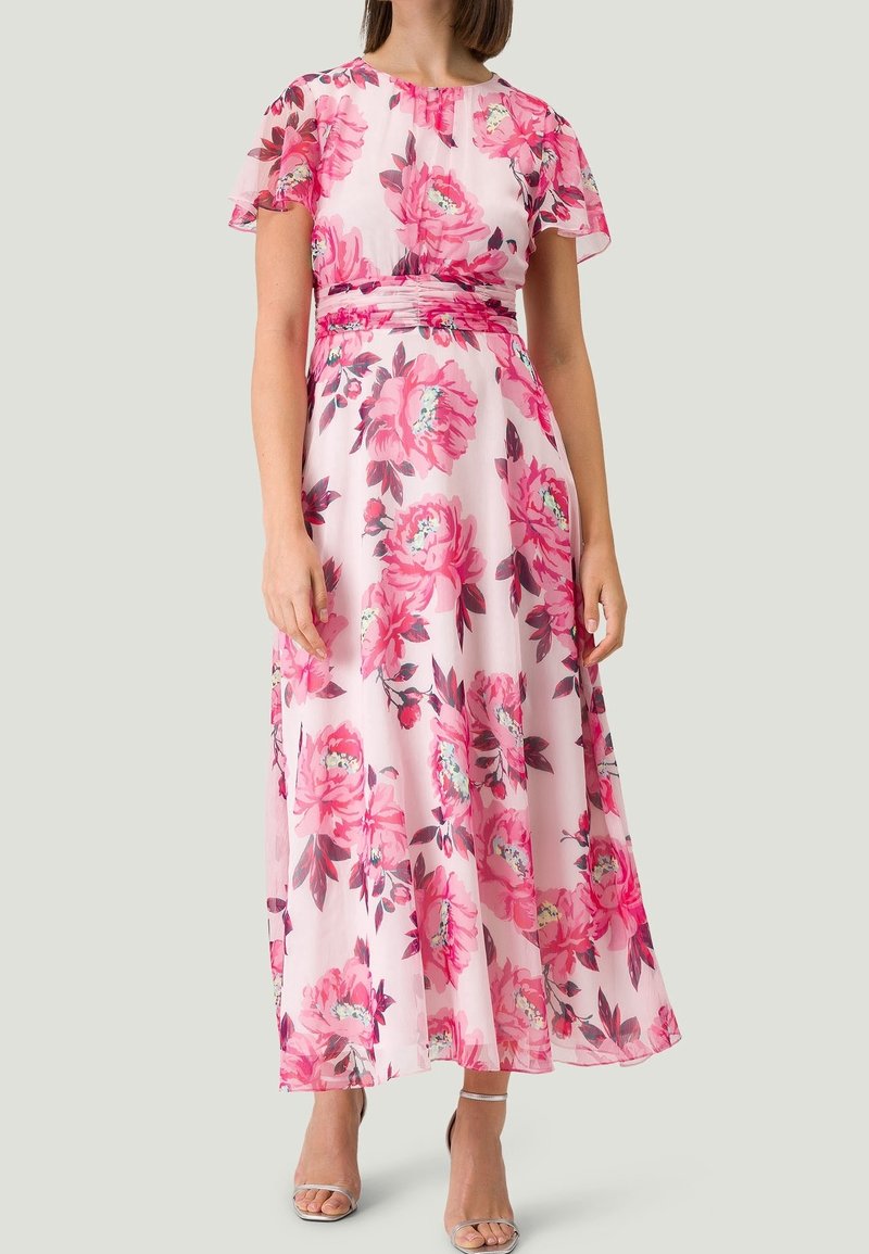 Femme portant une robe longue rose clair avec de grands motifs floraux rose vif et des manches volantées, associée à des sandales argentées à talons hauts.
