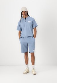Completo sportivo azzurro chiaro composto da una maglietta a maniche corte e pantaloni corti fino al ginocchio, realizzato in morbido tessuto con un logo a contrasto. Indossato con un cappellino beige.