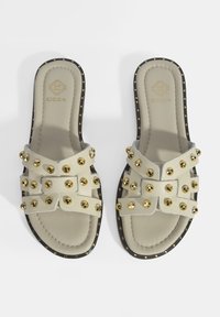 Beige leren slidersandalen met gouden studs, kruisstappen en een zwarte stippenrand rond de zool. Gevoelige voetbeddetails zichtbaar.