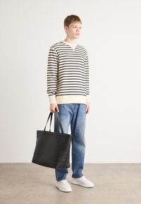 Stripet langermet skjorte i kremfarge og marineblå, kombinert med lyseblå jeans. Svart toteveske holdt i hånden, med glatt tekstur og robuste håndtak.