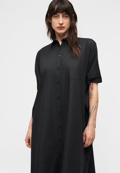 Άτομο με μακριά σκούρα μαλλιά φοράει ένα άνετο μαύρο φόρεμα με κουμπιά και κοντά μανίκια, που δείχνει τα τατουάζ στο αριστερό τους αντιβράχιο.