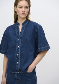 Camicia in denim a maniche corte, di colore blu e con colletto alla cinese. Presenta bottoni frontali e cuciture a contrasto lungo le cuciture.