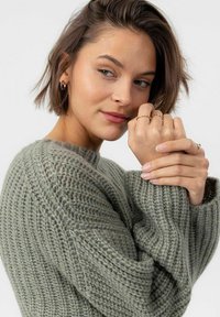 Maglione a maglia in verde tenue con un motivo testurizzato, caratterizzato da un ampio scollo e spalle scese. Mani decorate con anelli semplici.