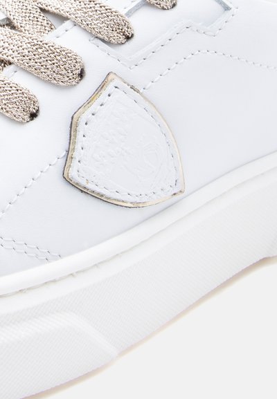 Λευκό δερμάτινο αθλητικό παπούτσι με μεταλλικά χρυσά στοιχεία, υφασμένα κορδόνια και λεπτομέρεια με λογότυπο θυρεού. Διαθέτει λεία επιφάνεια και λαστιχένια σόλα.