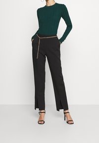 Groene geribbelde langemouwen top gecombineerd met zwarte broeken met zijsplitten, vastgemaakt met een gouden kettingriem. Zwarte sandalen met banden maken de look compleet.