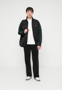 Sort lynlåstrøje med grønne striber, rød branding; hvid t-shirt; sorte bukser; holder en sort skuldertaske; hvide sneakers med brune detaljer.