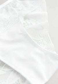 Bralette en dentelle blanche avec une base en tissu lisse, agrémentée d'accents délicats en dentelle et de motifs complexes le long des bords et des bonnets.
