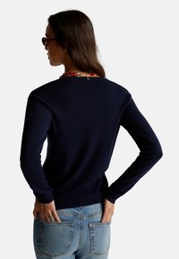 Námořnický modrý pletený svetr s kulatým výstřihem, pružnými rukávy a spodním lemem, v kombinaci se světle modrými džíny, které mají zadní kapsy.