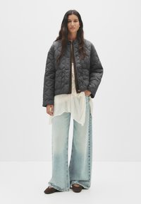 Vadderad grå jacka med rund halsringning, i kombination med en lager-på-lager krämfärgad topp och vida ljusblå jeans. Bruna skor fulländar outfiten.