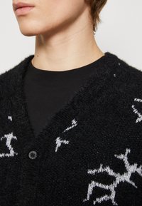 Juodas vilnonis kardiganas su V formos iškirpte, tekstūruotu mezgimu ir baltais snaigių raštais. Apima vieną juodą mygtuką krūtinėje.
