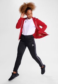 Rote Zip-Jacke, weißes T-Shirt, schwarze Hose mit seitlichem Chevron-Muster und schwarze Sportschuhe, Model posiert in einer aktiven Haltung.