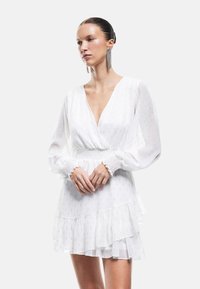 Witte jurk met lange, opgeblazen mouwen, diepe V-halslijn en een gelaagde, gerimpelde zoom. Textuurstof met subtiele metallic accenten. Gladde afwerking.
