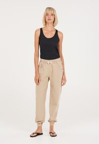 Tanktop aus dunkelgrauem Stoff, beige elastische Hosen mit Seitentaschen und schwarze Flip-Flops, vor einem schlichten hellen Hintergrund.