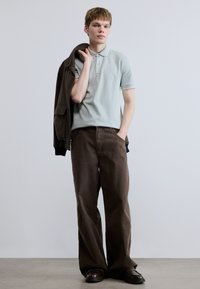 Ανοιχτό μπλε πόλο μπλουζάκι με κοντά μανίκια, καφέ παντελόνι με φαρδιά μπατζάκια και καφέ μπουφάν στον ώμο. Μαύρα παπούτσια ολοκληρώνουν την εμφάνιση.