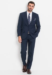 Completo blu navy con vestibilità strutturata, giacca monopetto, rever a scialle e pantaloni coordinati. Abbinato a una camicia bianca e una cravatta a righe.
