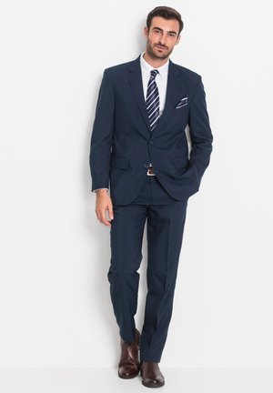 Marineblauer Anzug mit strukturiertem Schnitt, einreihiger Jacke, senkrechten Revers und passender Hose. Kombiniert mit einem weißen Hemd und einer gestreiften Krawatte.