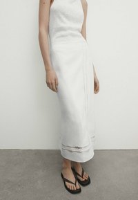 Biała lniana spódnica z koronkową lamówką, o prostej sylwetce. Połączona z białym, bezrękawnym topem i czarnymi sandałami.