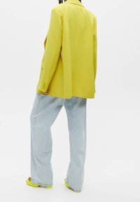 Personne portant un blazer jaune, un jean pattes d'éléphant bleu clair et des sandales jaunes, debout devant un fond blanc uni.