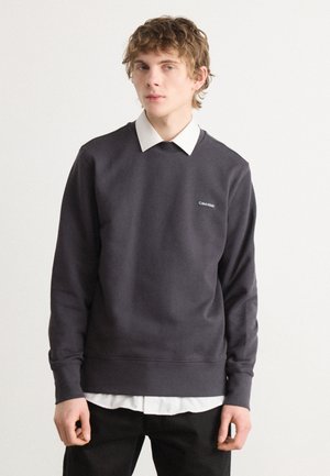 Mørkegrå sweatshirt lavet af bomuldsblanding, med rund halsudskæring og et lille hvidt logo på brystet, lagdelt over en hvid krave skjorte.