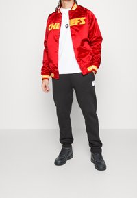 Giacca bomber rossa in satin con la scritta "CHIEFS" stampata in giallo, abbinata a una maglietta bianca e pantaloni della tuta neri. Sneakers nere completano l'outfit.