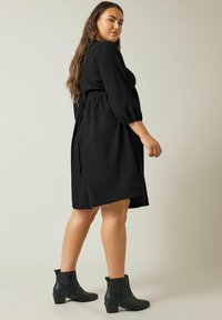 Schwarzes knielanges Kleid aus strukturiertem Stoff, mit Rundhalsausschnitt, Dreiviertelärmeln und gebundenem Taillengürtel. Trägt dazu schwarze Knöchelstiefel.