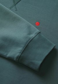 Dunkelblauer Sweatshirt mit strukturiertem Bündchen, einem auffälligen roten Stickpunkt und glattem Stoff. Die Details umfassen kontrastierende Nähte.