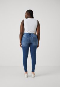 Skinny jeans in medium blue denim, featuring a high waist, back pockets, and subtle whiskering. Paired with a white sleeveless top.