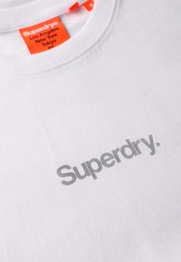 Camiseta de algodón blanca con escote de cuello redondo y un logotipo gris "Superdry" en la parte delantera. Etiqueta naranja brillante en el cuello con detalles de la marca.