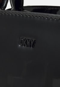 Svart handväska i patentläder med präglad DKNY-logotyp på framsidan, med en slät yta och subtil glans. Handtagen är rundade och stabila.