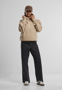 Beige hettegenser med en frontlomme, kombinert med mørke bukser med sidesømmer og subtle detaljer. Brun caps fullfører antrekket.