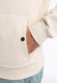 Hellbeige Sweatshirt mit lockerer Passform, ausgestattet mit einer großen frontalen Tasche und schwarzen Druckknopfdetails. Gerippte Bündchen betonen die Ärmel.