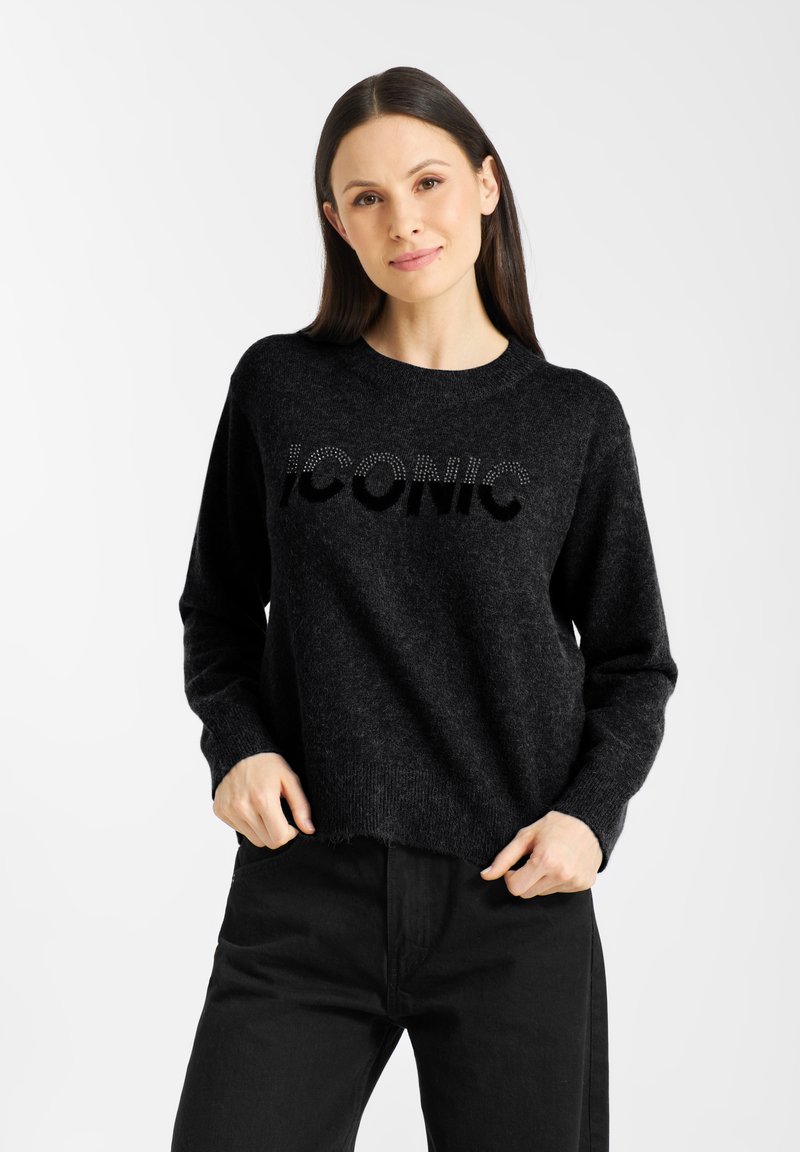 Zwarte gebreide trui met "ICONIC" in textuur letters. Heeft een geribde ronde hals en manchetten, en een cropped zoom. G gedragen met zwarte broek.