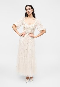 Femme debout, mains sur les hanches, portant une robe blanche brodée de fleurs, à manches bouffantes et longueur cheville, avec des escarpins blancs pointus.