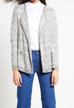Kobieta ubrana w szary, wzorzysty kardigan z rozpiętym przodem na białej koszuli zapinanej na guziki oraz niebieskich dżinsach na jednolitym białym tle.